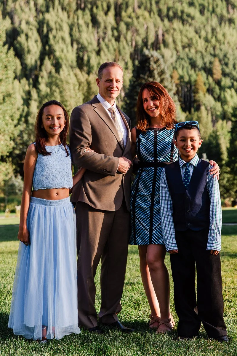 Family of four smiling in a scenic outdoor setting. The parents are dressed formally; the daughter in a blue dress, son in a vest. Green forest backdrop.
