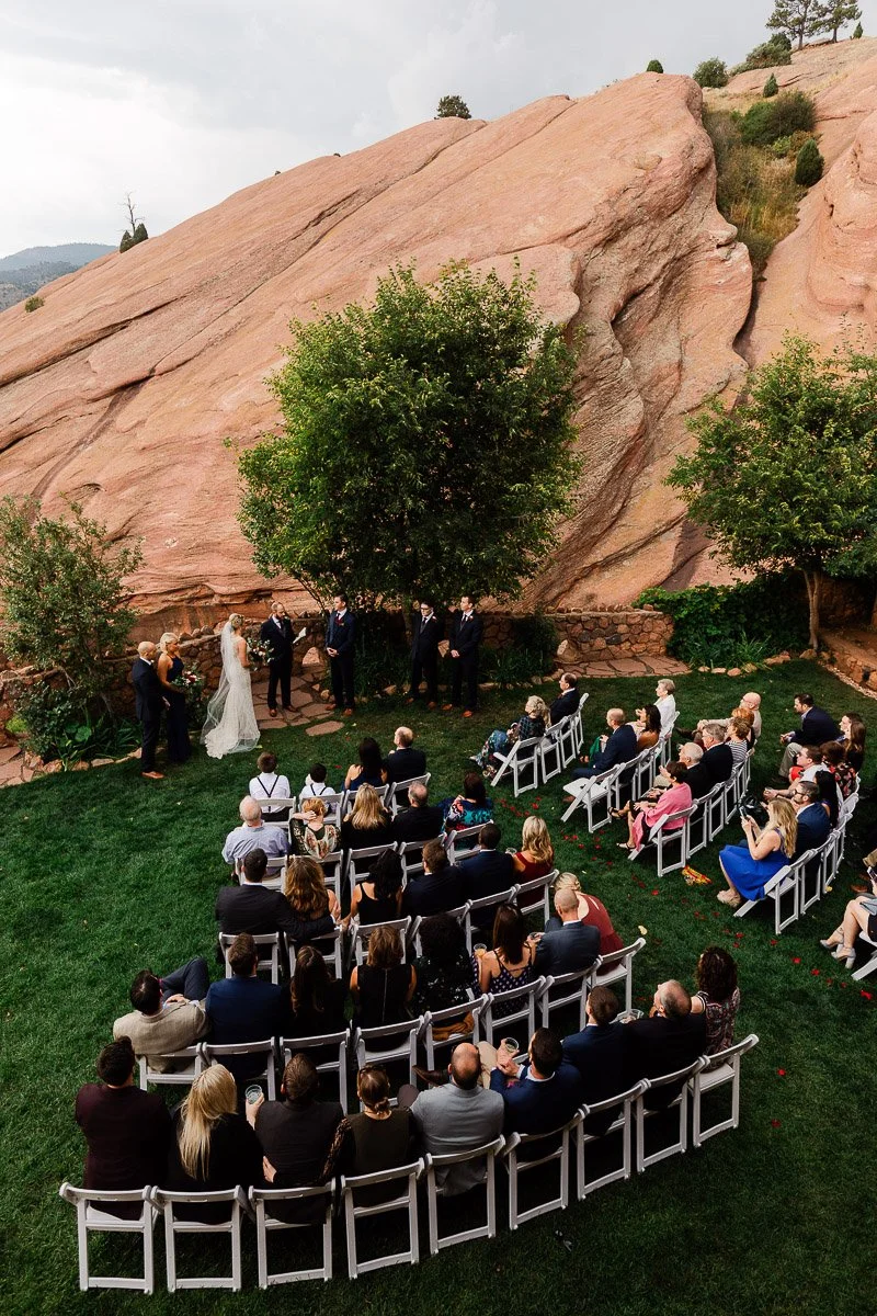 Wedding ceremony outdoors with guests seated on a green lawn. A couple stands at the altar under trees, with red rock formations in the background. Serene and intimate.
