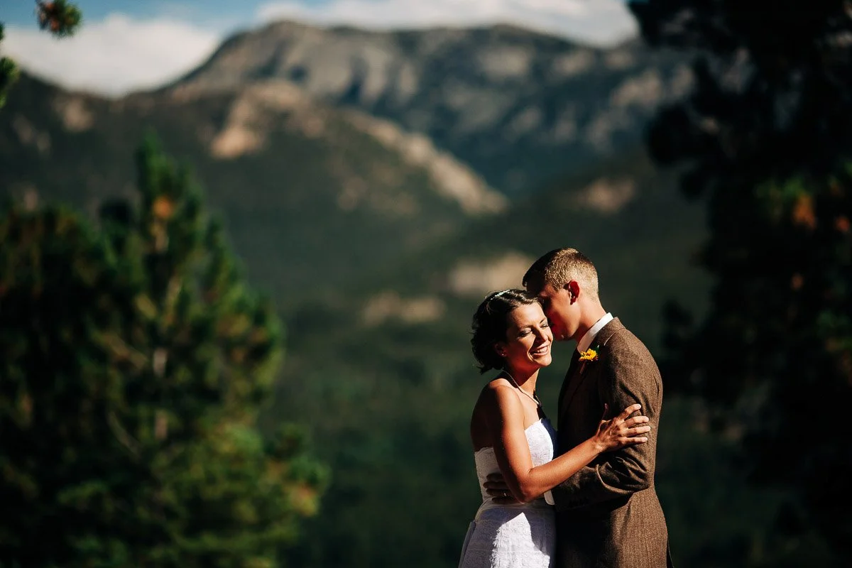 A couple, dressed in wedding attire, embrace and smile against a backdrop of majestic mountains and trees. The scene conveys romance and joy captured by YMCA Of the Rockies wedding photographer tomKphoto