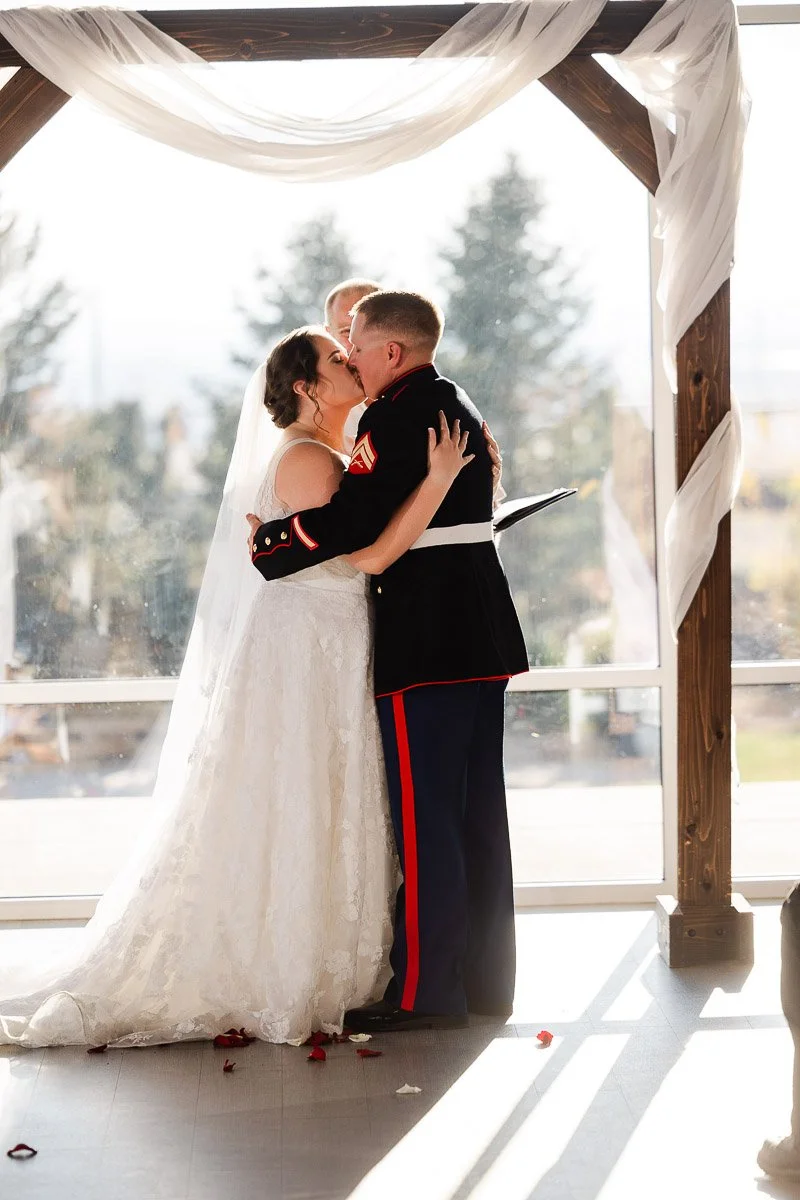 A bride in a lace gown and a groom in a military uniform share a kiss under an arch draped with white fabric, set against a sunny outdoor backdrop.