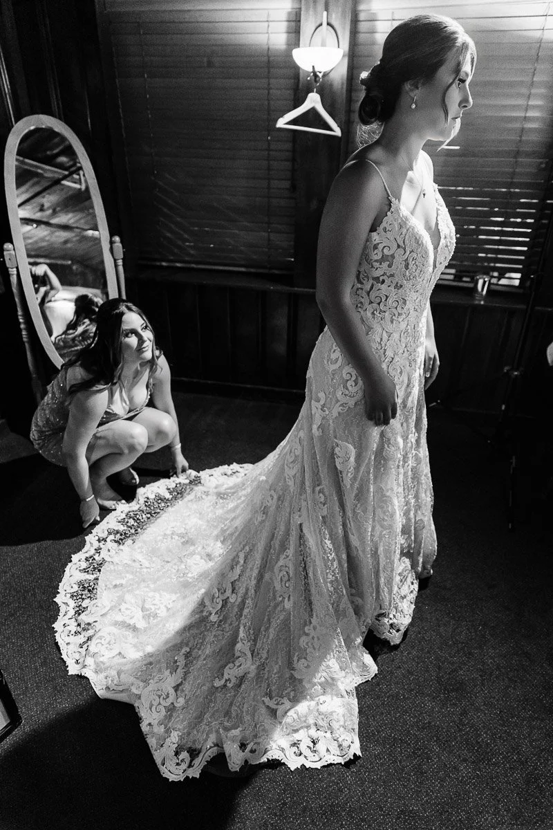 A bride in a lace dress stands in a dimly lit room, as an attendant adjusts her train. A standing mirror reflects part of the scene, adding elegance.
