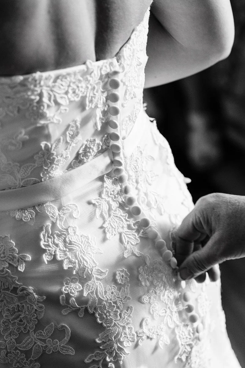 Close-up of hands buttoning a lace wedding dress, emphasizing delicate floral patterns. The black-and-white image conveys a sense of anticipation and elegance.