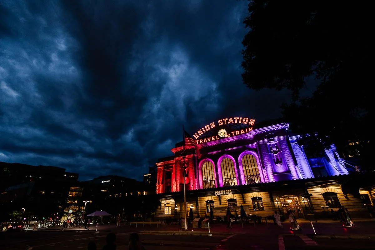 Dramatic evening view of a train station lit in red and blue. The sky is dark and cloudy, enhancing the vibrant colors and creating a striking contrast.