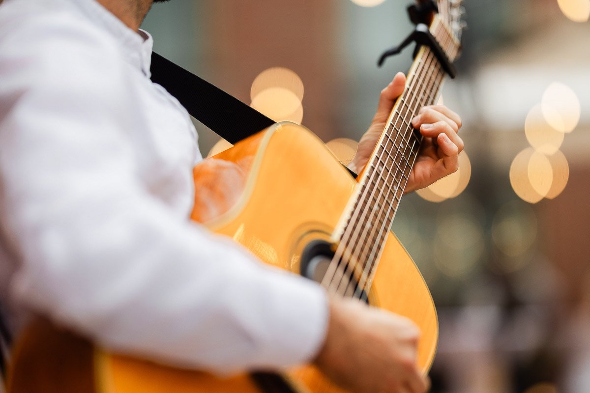 A person plays an acoustic guitar, wearing a white shirt, with blurred bokeh lights in the background, creating a warm and relaxed atmosphere.