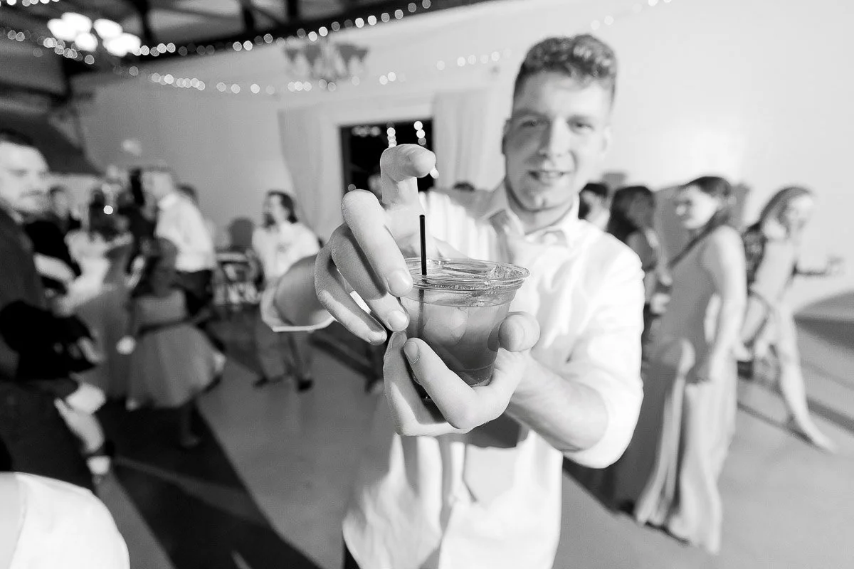 A man in a white shirt offers a drink with a straw at a party. People are dancing in the blurred background, and festive lights hang above.