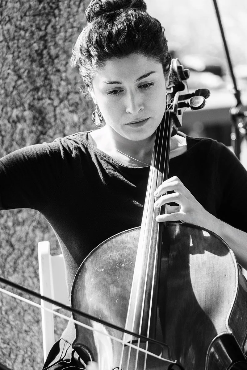 Black and white image of a focused woman playing a cello outdoors. She has a serious expression, with her hair in a bun, wearing a dark top.