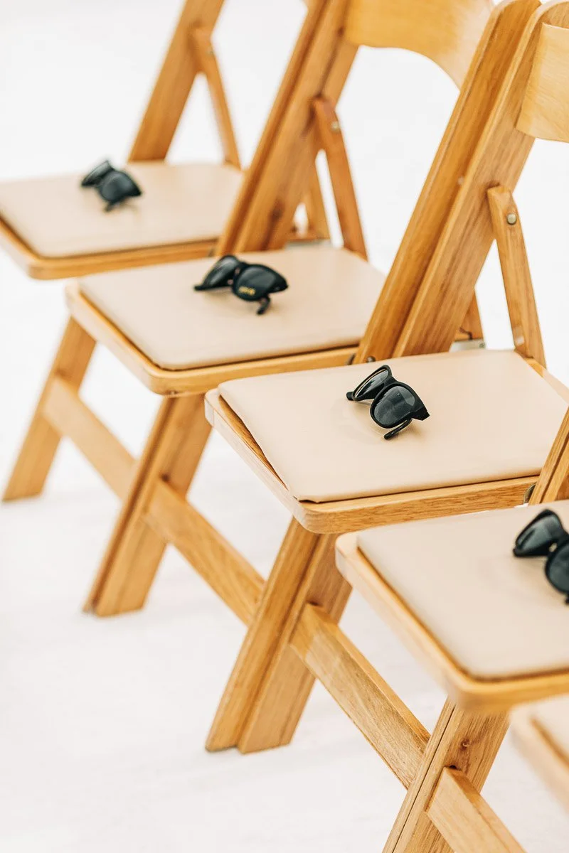 Close-up of three wooden folding chairs with beige cushions in a row on snow. Each chair has a pair of sunglasses on the seat, evoking a casual, wintery vibe.
