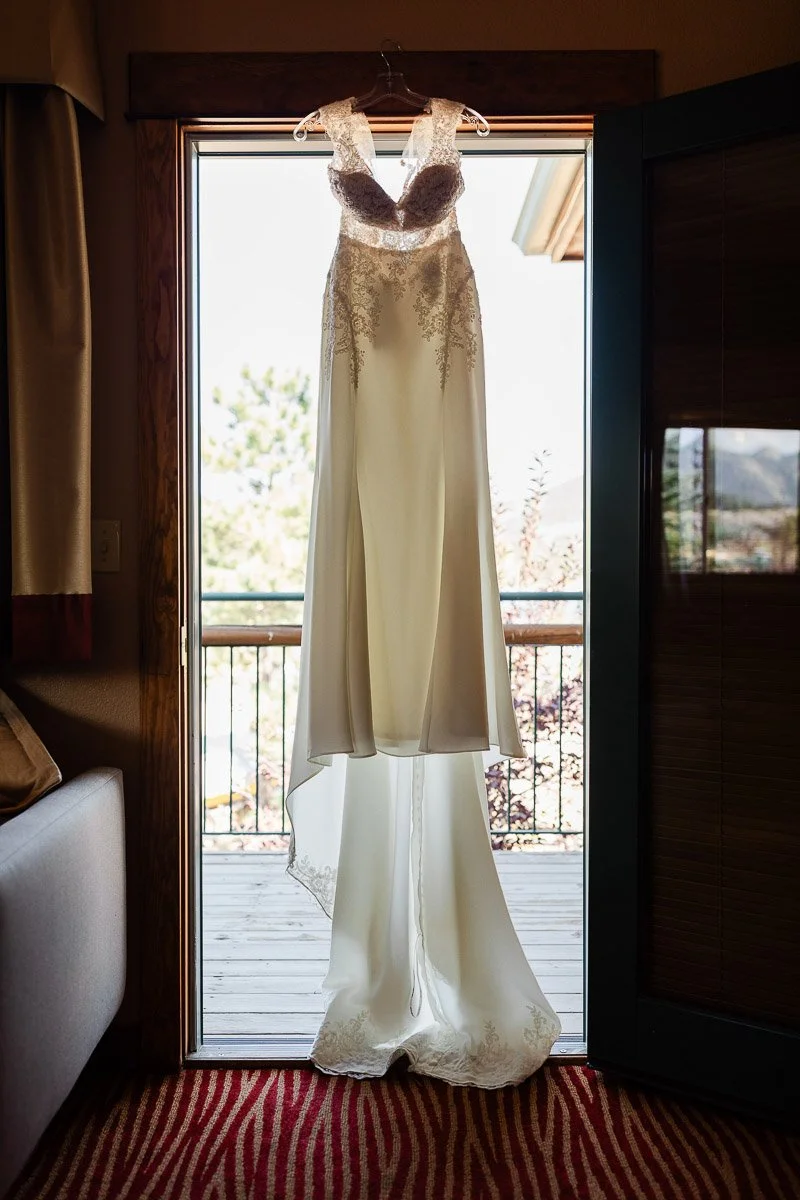 Elegant white wedding dress with lace bodice hangs in a sunlit doorway, revealing a balcony with soft, natural scenery. The mood is serene and romantic.