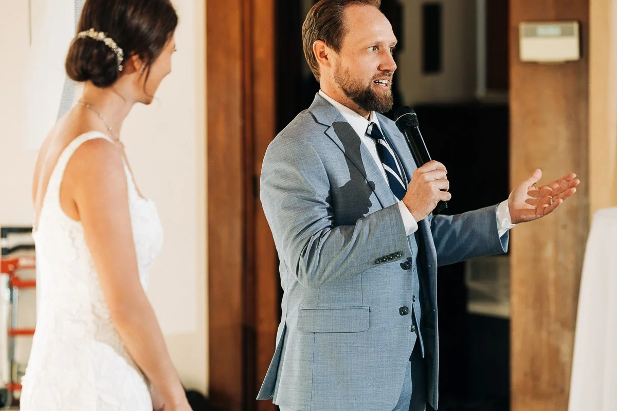 A man in a light blue suit speaks into a microphone, gesturing with one hand, while a woman in a white dress listens intently. The scene conveys a joyful and celebratory mood, likely at a wedding.