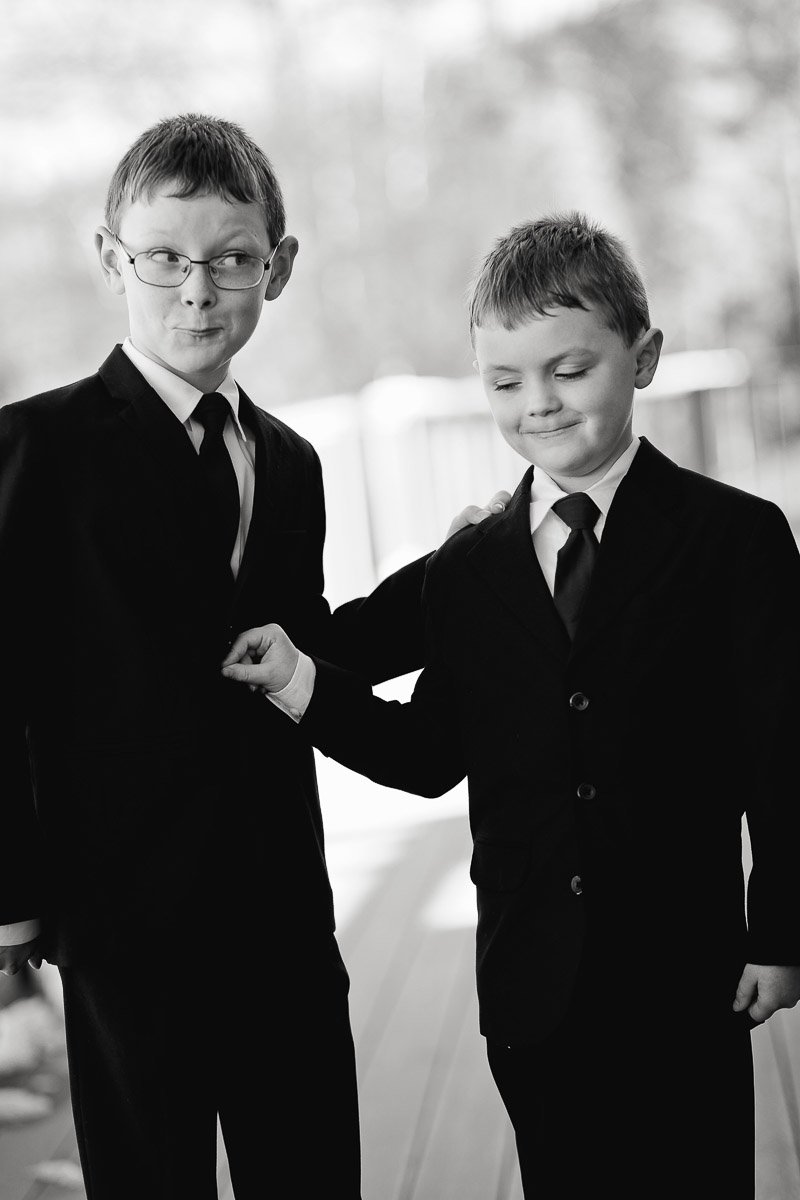 Two young boys in suits and ties stand side by side. One boy makes a playful face, while the other smiles softly. The tone is lighthearted.