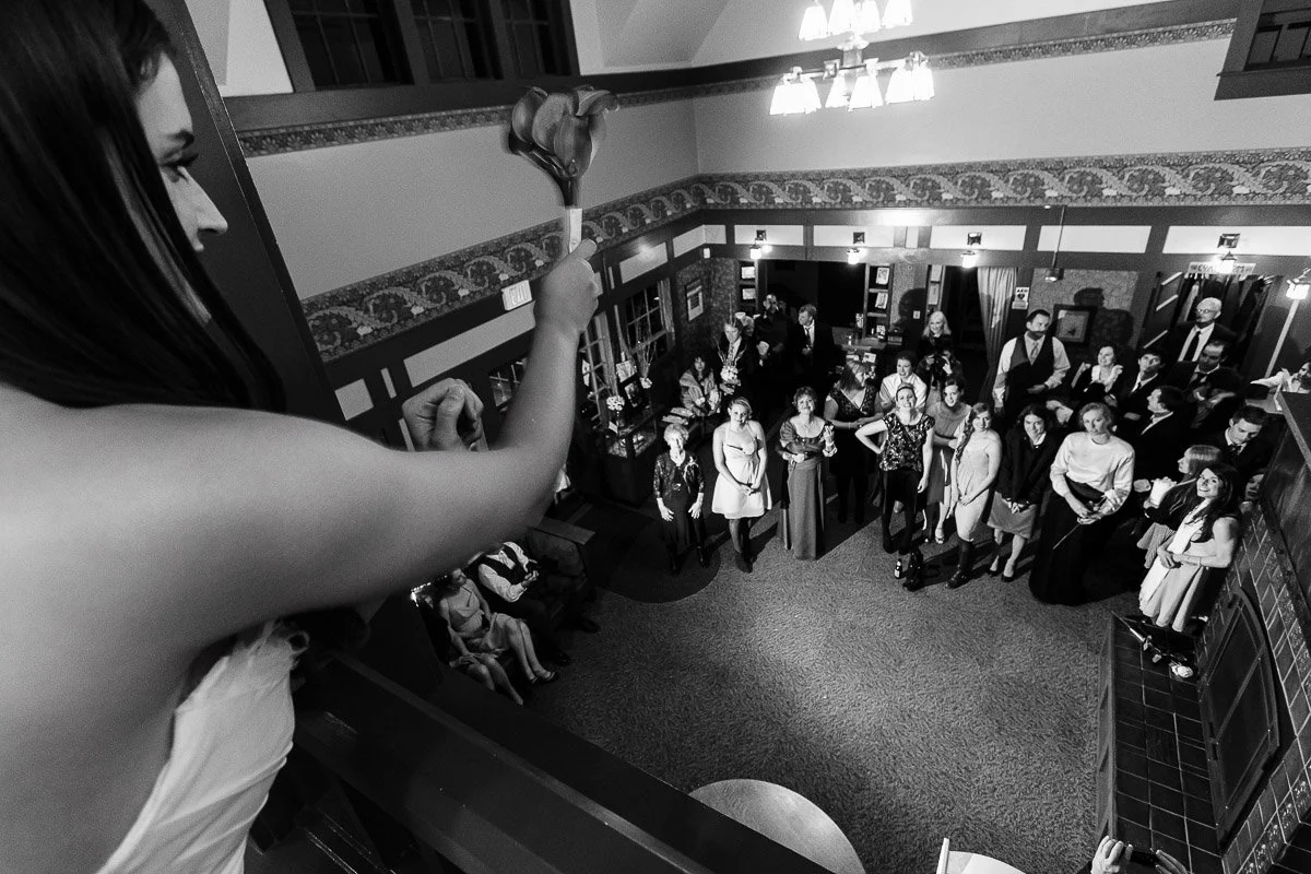 A woman in a strapless dress holds flowers, preparing to toss them to a group of smiling people below in an elegantly decorated room. Black and white image.