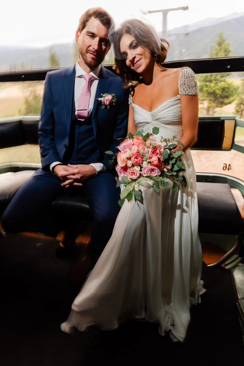 A newlywed couple sits in a gondola, with the bride in a white gown holding a pink rose bouquet. The groom is in a navy suit with a pink tie. Natural light and trees create a warm, serene atmosphere.