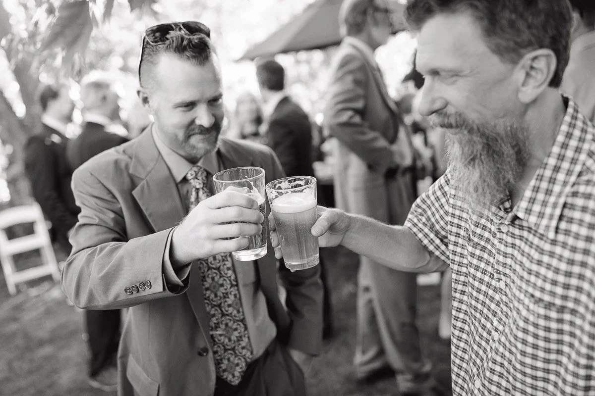 Two men cheer with beer glasses at a casual outdoor gathering. One wears a suit, the other a checkered shirt. They appear joyful and relaxed.