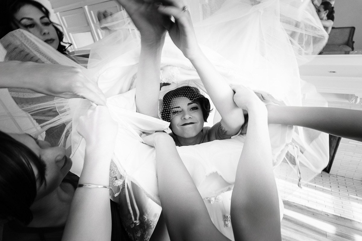 Black and white photo of a joyful bride being assisted into her dress, surrounded by helping hands and tulle layers, capturing a moment of excitement.