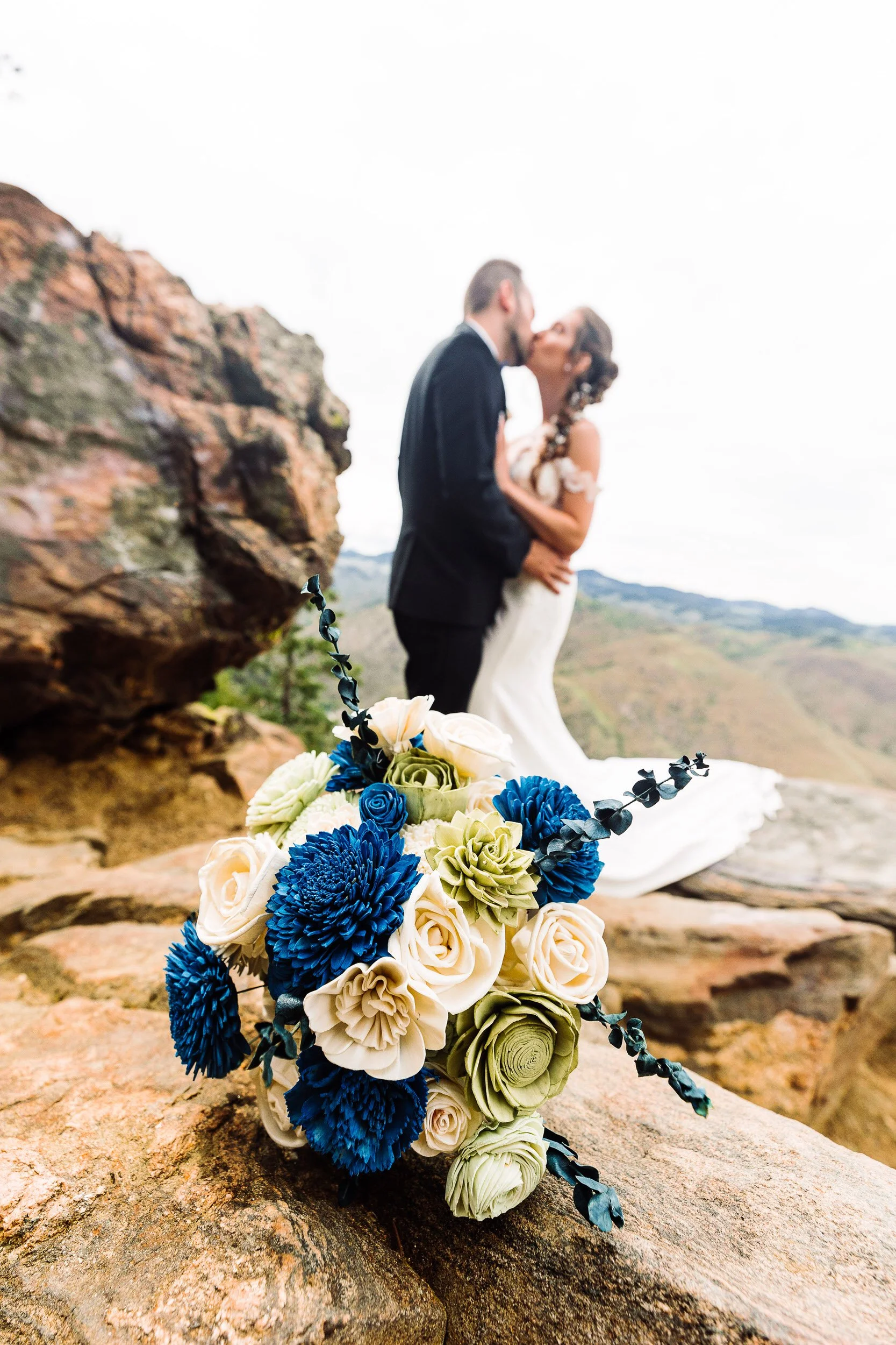 Bride's bridal boquet rests on a rock while wedding couple kisses in background soft focus at Mount Vernon Canyon Club in Golden, Colorado.