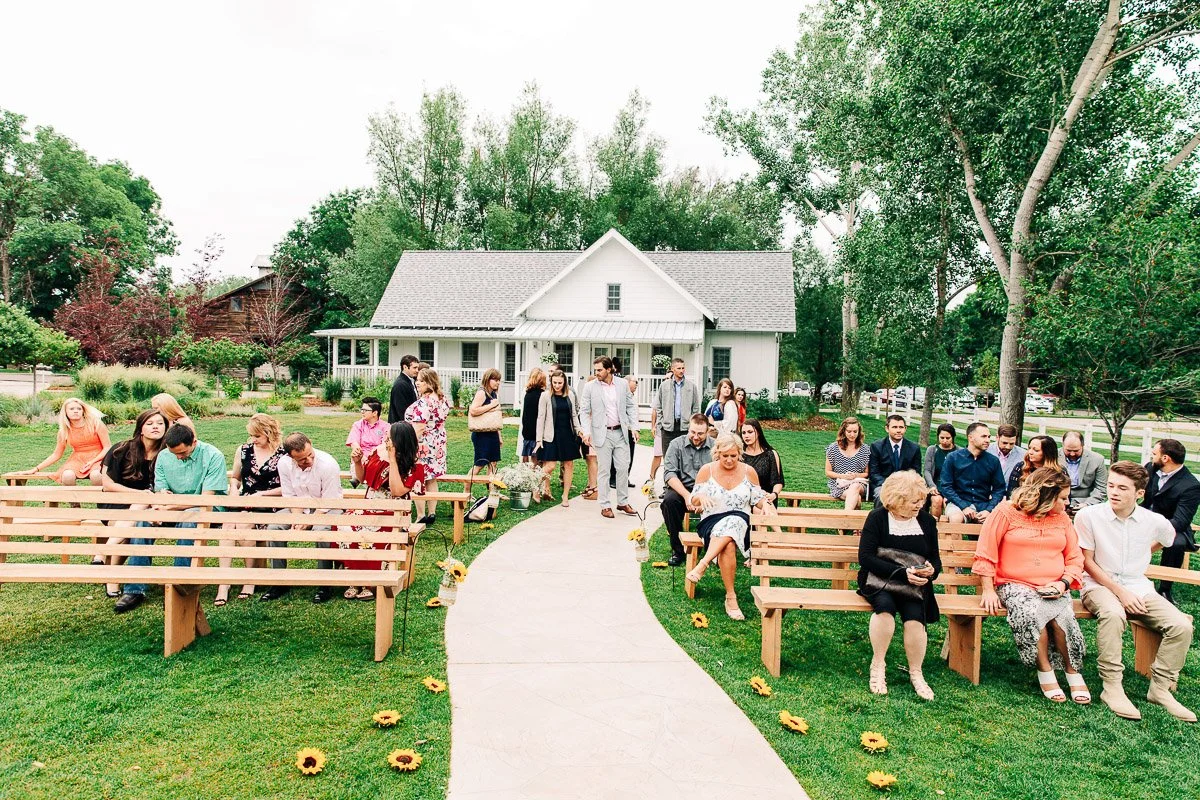 Guests sit on wooden benches in a garden wedding setting, chatting and smiling. A path lined with sunflowers leads to a white house, creating a joyful, relaxed atmosphere.