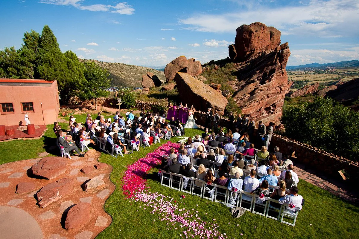 Aerial view of an outdoor wedding in a rocky landscape. Guests sit on white chairs with a pink petal aisle. Red rocks and blue sky enhance the lively scene.