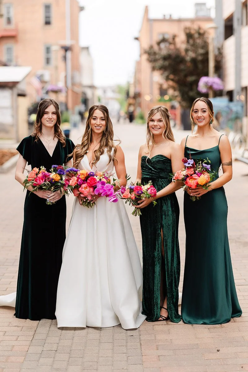 Bridal party standing on a brick path in a city, with a bride in a white gown and three bridesmaids in green dresses holding vibrant bouquets.