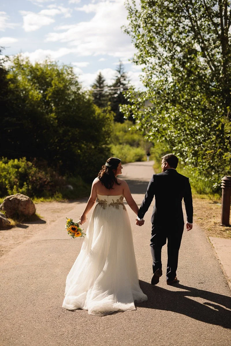 A bride in a white gown and a groom in a dark suit walk hand-in-hand down a sunlit path, surrounded by lush greenery and under a partly cloudy sky.