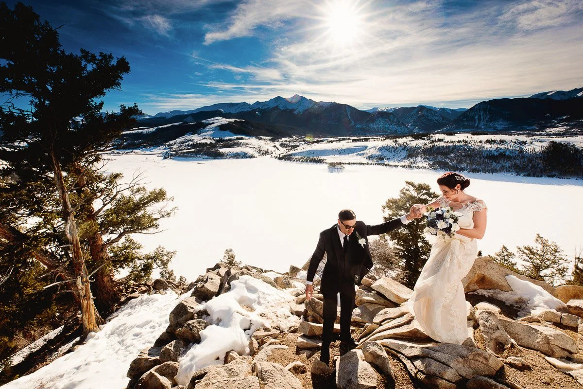 A bride and groom in wedding attire stand on snow-covered rocks, with a vast snowy landscape and mountains under a bright, sunny sky in the background.