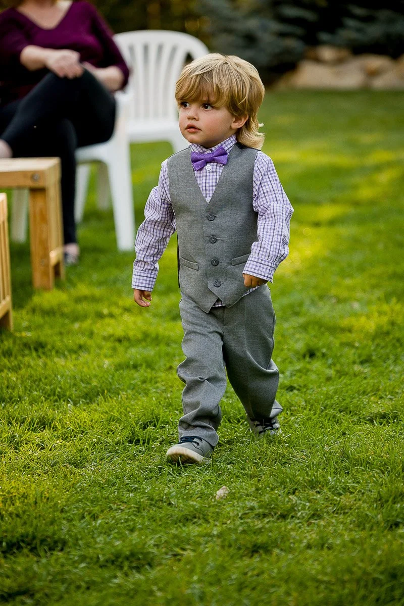 A young boy with blonde hair walks confidently on grass, wearing a gray suit with a purple bow tie. A white chair and seated person are blurred in the background.
