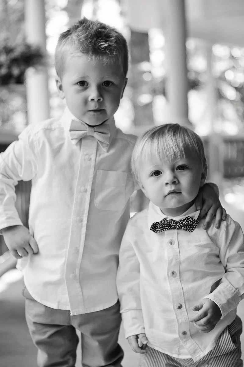Two young boys stand close together, both in white shirts and bow ties. The older boy has his arm around the younger, showing a sweet bond.