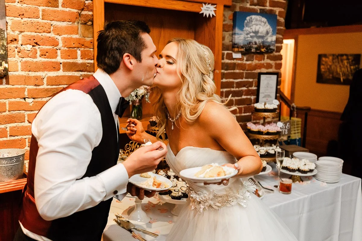 A bride and groom kiss joyfully, holding plates of cake, against a cozy brick wall backdrop next to a cake table adorned with cupcakes and flowers.