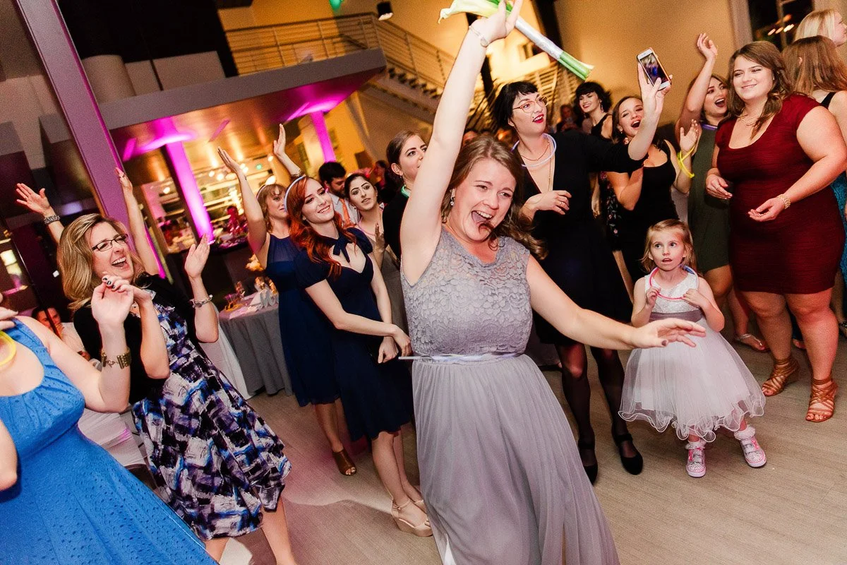 A joyful group of women dancing and celebrating indoors with vibrant pink lighting. A woman in the foreground smiles broadly, raising her hand in excitement.