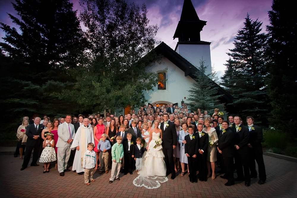 A large wedding party gathers in front of a quaint church at dusk, under a purple-hued sky. The bride and groom stand at the center, surrounded by guests captured by Colorado Wedding Photographer tomKphoto