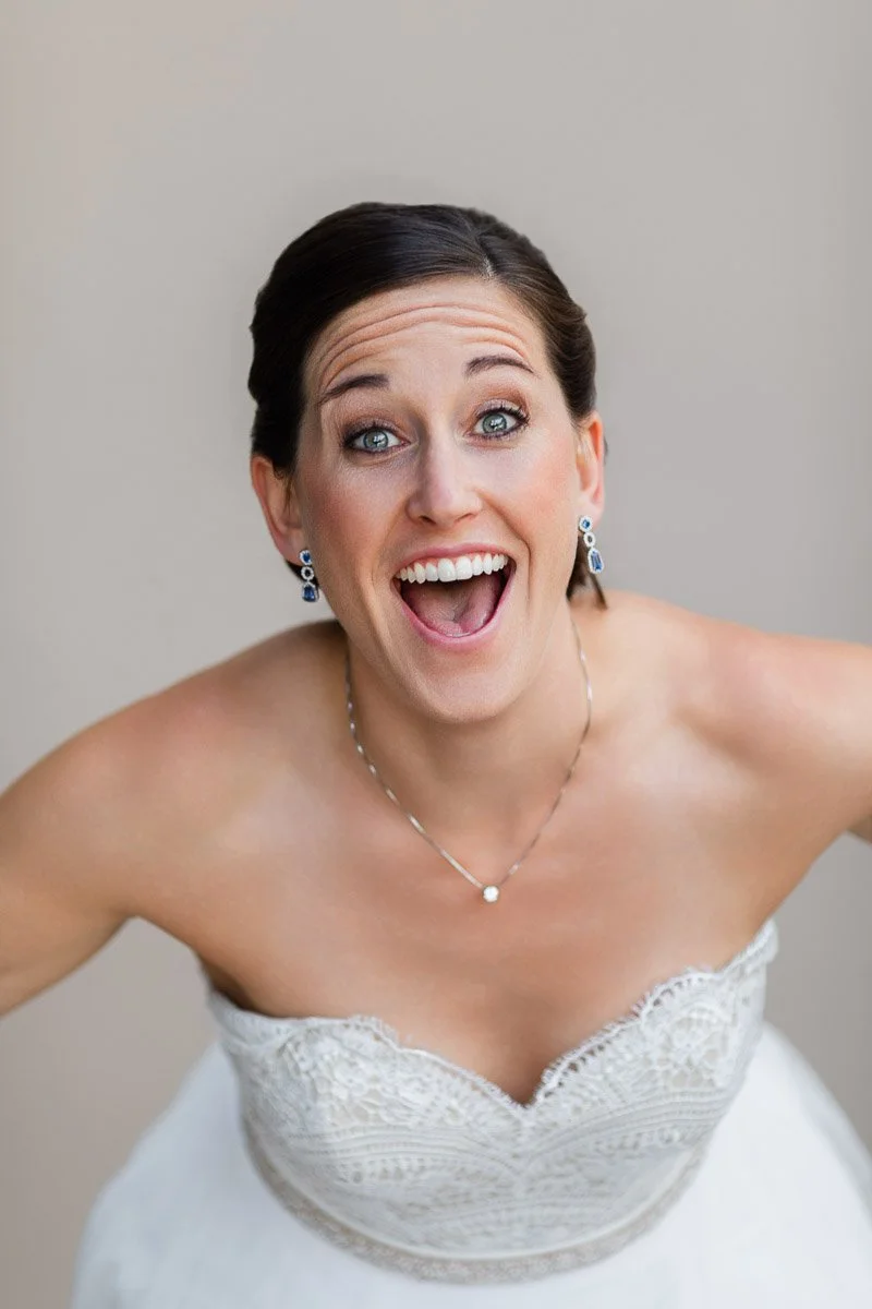 Bride laughing joyfully, leaning forward in a strapless lace wedding dress, with elegant jewelry. Neutral background enhances her happy expression.