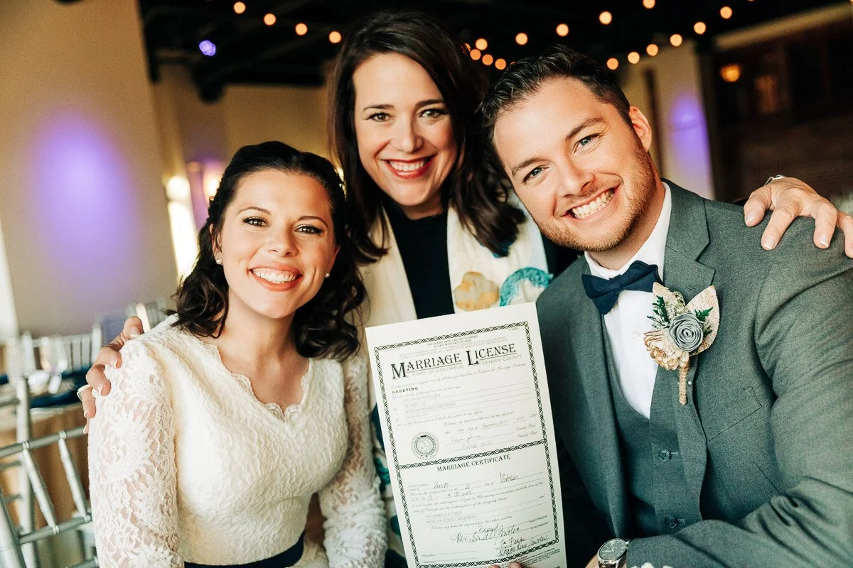 Smiling couple in wedding attire holds a marriage license, with a woman standing behind them, in a warmly lit venue decorated with string lights.