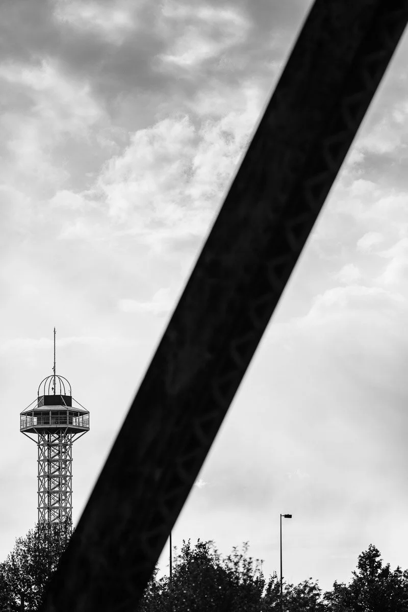 Black and white image of a tall observation tower with a geometrical structure in the foreground. Overcast sky and trees add a serene tone.