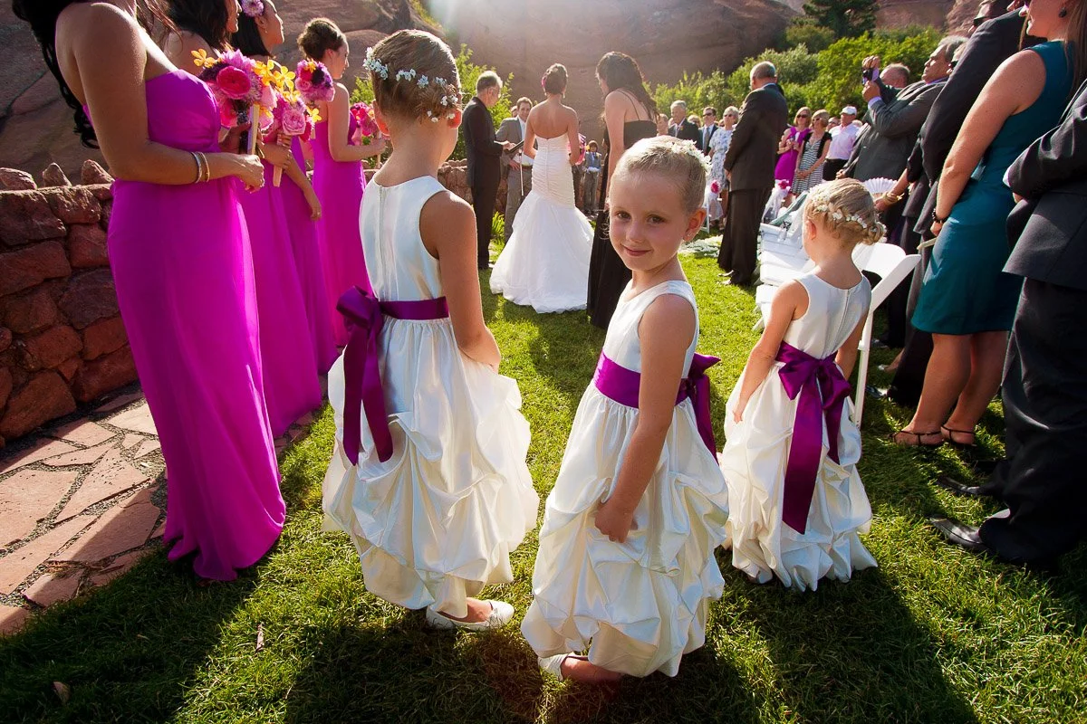 Three young flower girls in white dresses with purple sashes stand on a sunny lawn during an outdoor wedding. Guests and bridesmaids in pink look on.