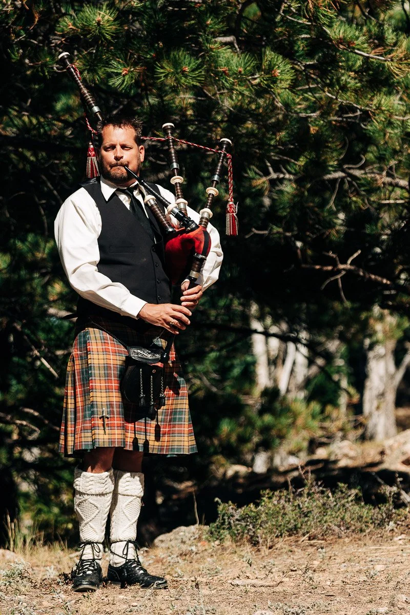 A man in a traditional Scottish kilt and vest plays bagpipes outdoors. He stands on earthy terrain, surrounded by lush green pine trees, conveying a serene, rustic atmosphere.