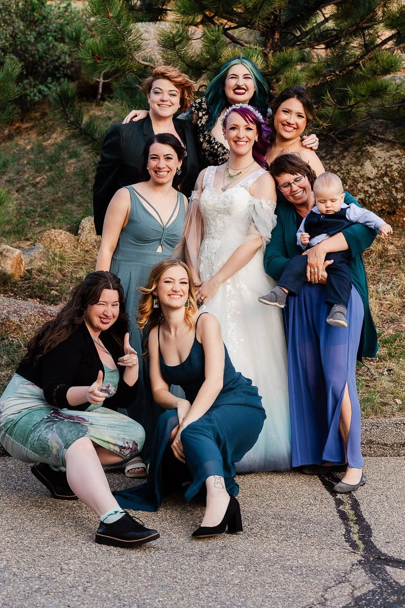 A joyful group photo outdoors with eight women in formal attire, smiling. The bride in white is central, holding hands with friends, one holding a baby.