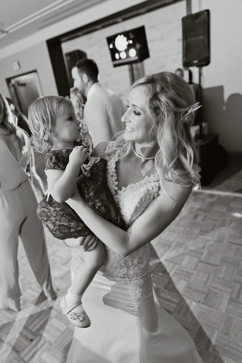 A joyful woman in a lacy dress holds a smiling toddler on a dance floor. Others dance in the background, creating a lively celebration atmosphere.