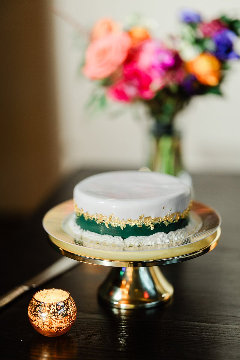 A small round cake with white icing and gold accents sits on a gold stand. A vibrant bouquet with pink and orange flowers is blurred in the background.