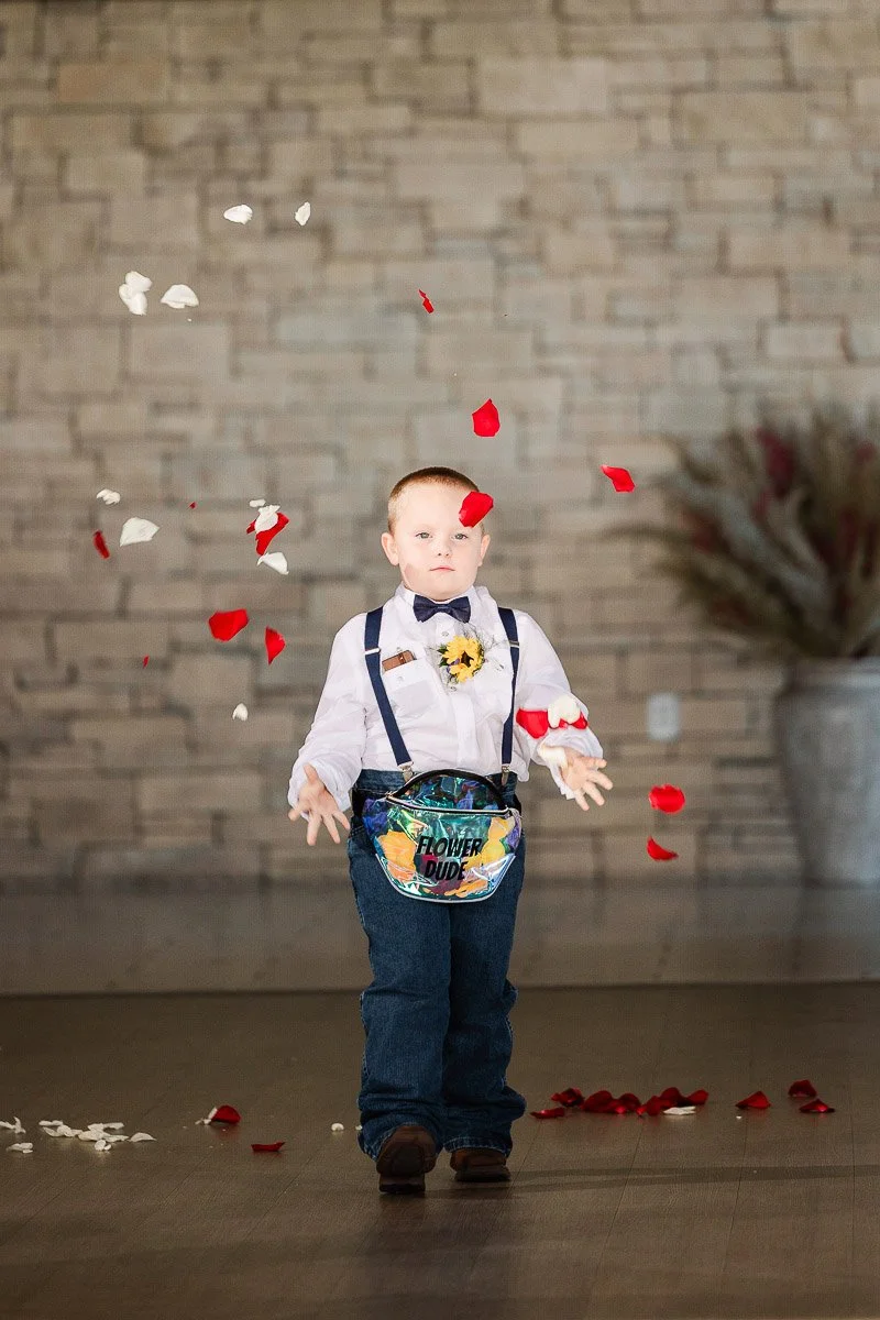 A young boy dressed in a bow tie and suspenders joyfully tosses red and white petals, standing in a rustic setting. The mood is playful and festive.
