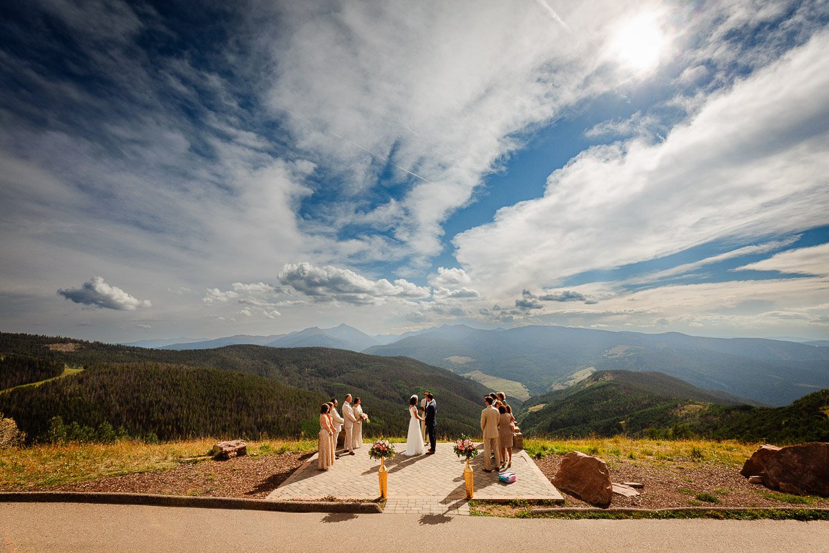 A couple weds on a scenic mountain platform under a vast, dramatic sky. Bridesmaids and groomsmen stand nearby, enhancing the serene, joyous atmosphere.