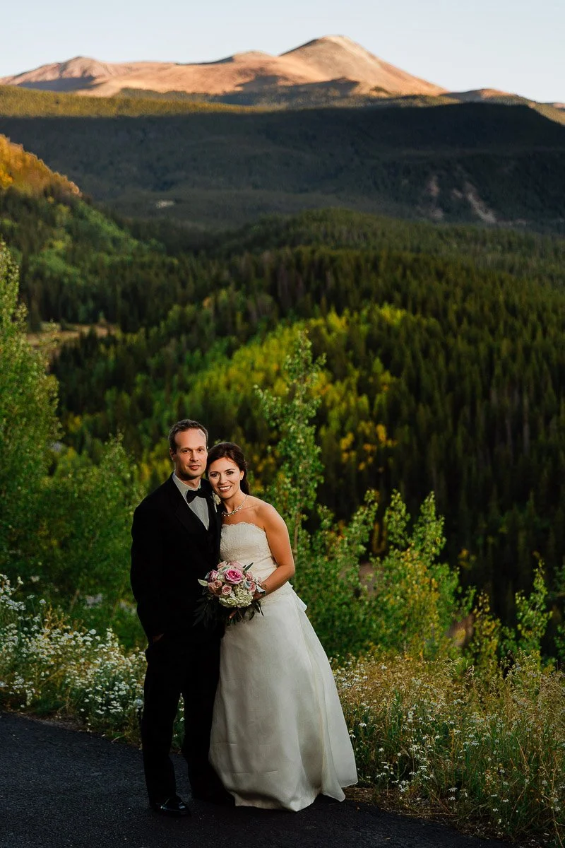 Bride and groom stand on a mountain path, surrounded by lush green trees and wildflowers, with sunlit peaks in the background, exuding romance and serenity.