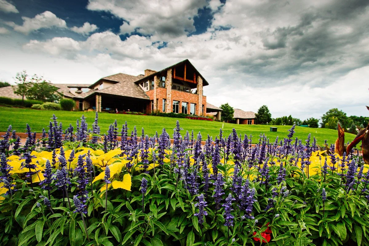 Beautiful brick Lakewood Country Club house surrounded by lush green lawn and vibrant purple and yellow flowers under a dramatic cloudy sky, creating a serene mood.