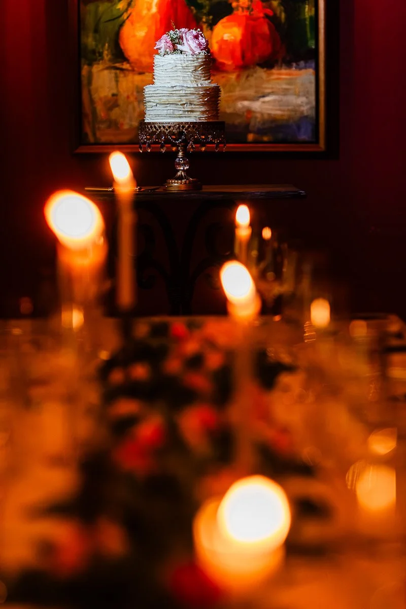 A tiered cake with pink flowers sits on an ornate stand against a vibrant painting backdrop. In the foreground, soft-focus candles create a warm ambiance.