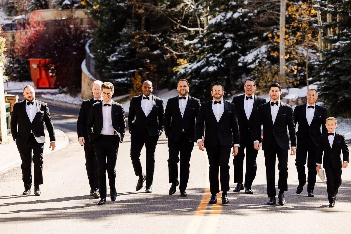 A group of men dressed in tuxedos, including a young boy, walk confidently down a sunlit road surrounded by snow-dusted trees and colorful foliage.