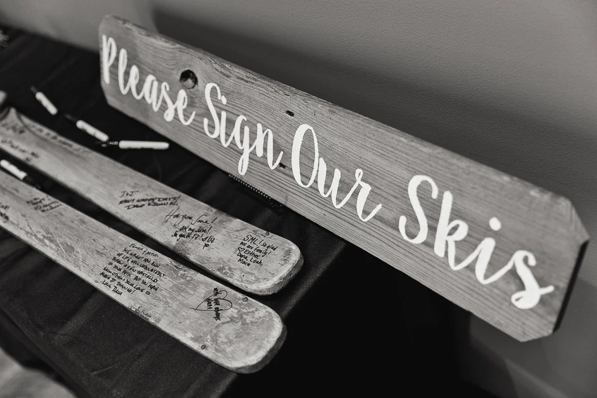 Black and white image of a wooden sign reading "Please Sign Our Skis" next to vintage skis covered in handwritten messages on a table. Nostalgic atmosphere.
