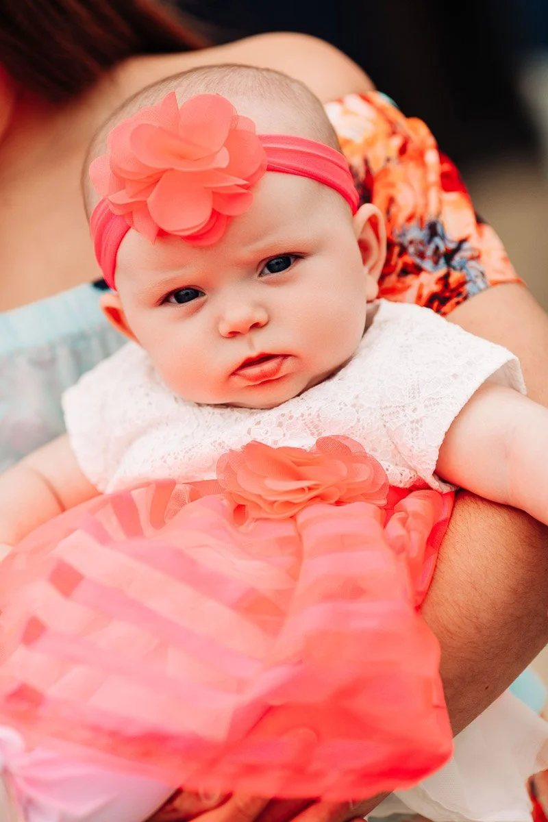 A baby with a serious expression is held in arms, wearing a pink headband with a large flower, a lace dress, and a bright pink tulle skirt.