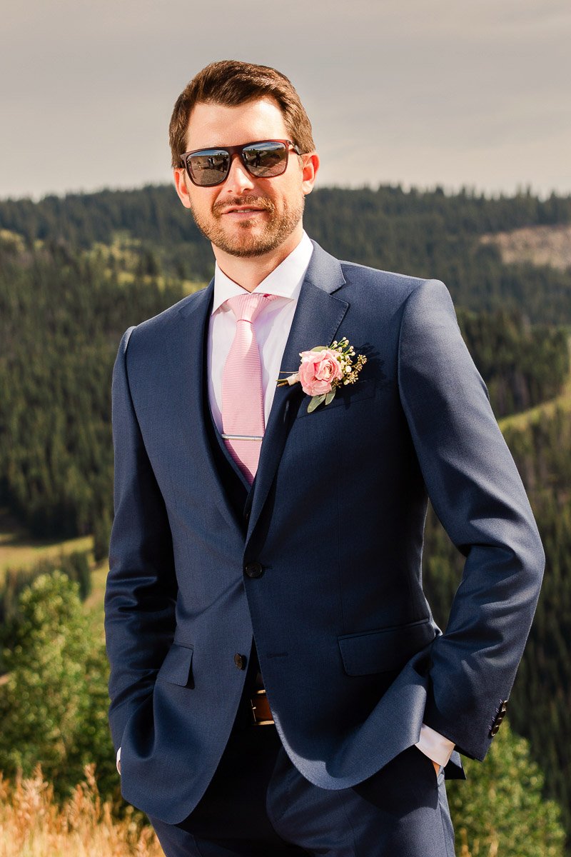 Man in a blue suit with a pink tie and rose boutonniere stands confidently outdoors. He wears sunglasses, with forested hills in the background.