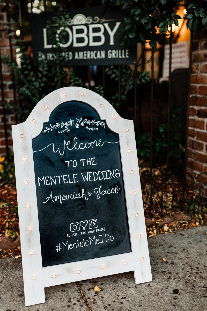 Chalkboard sign with lights welcomes guests to Mentele Wedding in front of rustic venue. Includes hashtag and social media icons for photo sharing.