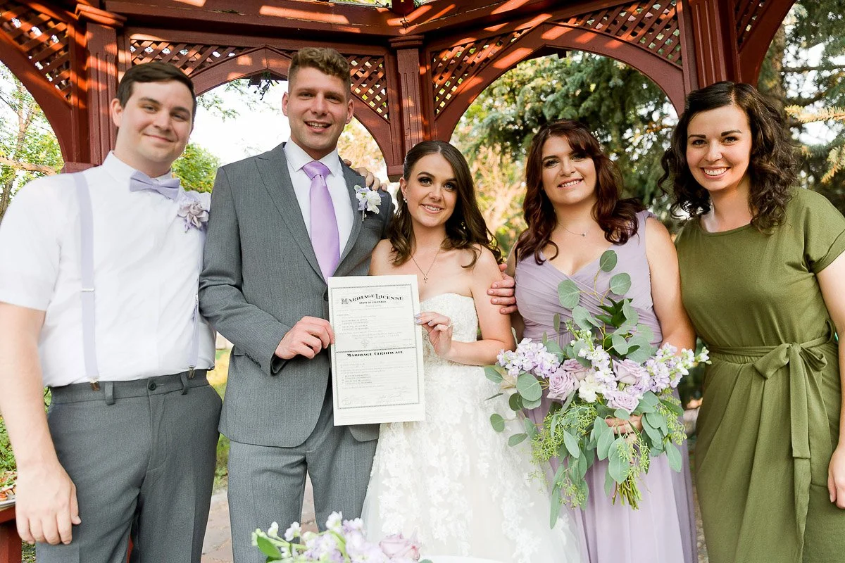 A couple stands joyfully with three people under a gazebo, holding a marriage license. The bride wears a white dress, and two women hold lilac bouquets.