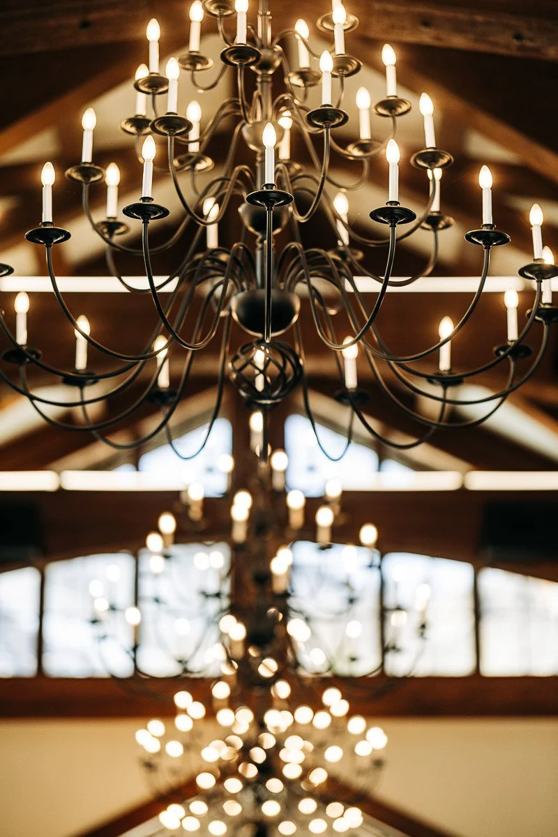 Grand chandelier with glowing candles in a dimly lit room with wooden beams. Soft, warm lighting creates an elegant and inviting atmosphere.
