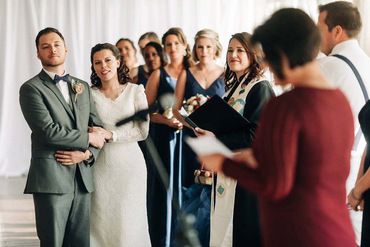 A bride and groom stand together in a wedding ceremony, surrounded by the bridal party. An officiant with a book is speaking, conveying a joyful, formal tone.